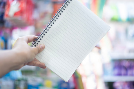 A person's hand is holding open a spiral-bound notebook with lined pages. The notebook is held at a slight angle, displaying the ruled sheets ready for writing. The background is softly blurred, showing the bright and colorful shelves of a retail store aisle stocked with various products. The focus is sharp on the notebook and the hand, emphasizing the potential for notes, ideas, or planning. The lighting is bright and even, creating a clear and usable image for concepts related to education, organization, and creativityの写真素材