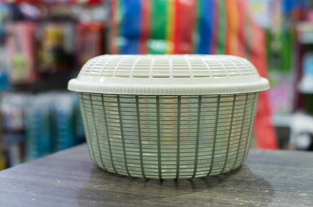 A head-on shot of a small, lidded plastic basket sitting on a dark wooden table. The basket is a pale green or off-white color with a distinctive woven or lattice pattern and a smooth, removable lid. The background is a blurry but colorful retail setting, with shelves of various products and a striped pattern, suggesting the photo was taken in a store or market. The focus is sharp on the basket, highlighting its texture and design, while the soft background provides context without being distractingの写真素材