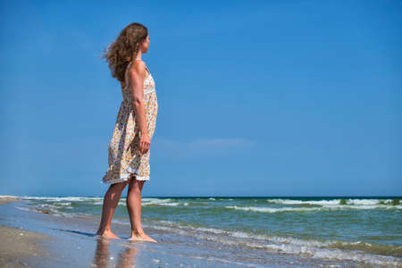 Young girl in a flower sundress stands on the seashore and looks into the distanceの写真素材