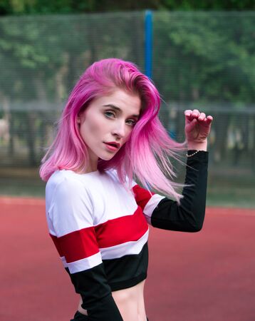 Portrait slim sensual girl cheerleader with pink hair and blue eyes in sportswear posing outdoors near fence at stadiumの写真素材