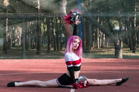 Young woman cheerleader with pink hair sitting in twine and holding red silver pompoms at stadiumの写真素材