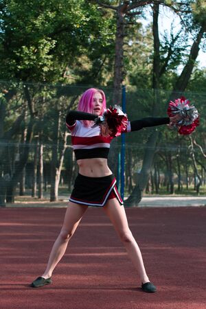 Slim fit girl cheerleader with pink hair dancing outdoors with red and silver pompoms at the stadiumの写真素材