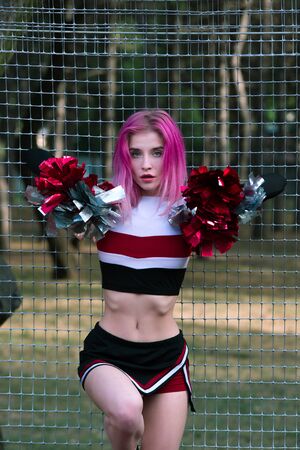 Slim fit girl cheerleader with pink hair in sportswear posing outdoors near fence at stadium with...