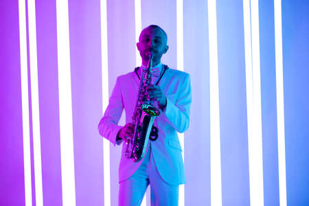 Happy saxophonist plays music on sax in elegant white suit. Man stand in the studio against the backdrop of neon lamps with blue backlight.の写真素材