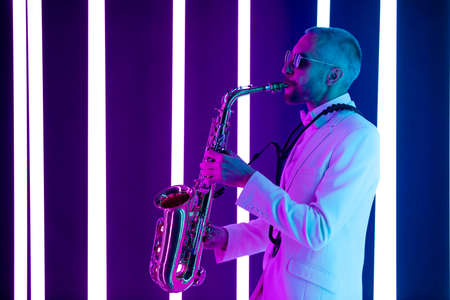 Side view stylish man in a white suit with a bow tie plays the saxophone in the studio. Saxophonist against the background of neon lamps with blue backlight.の写真素材