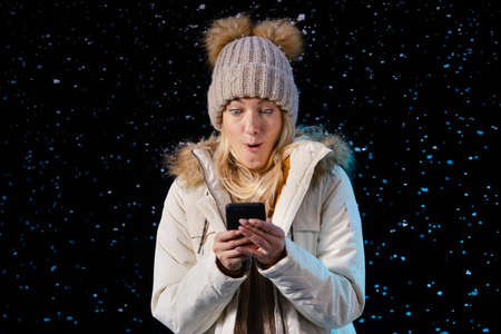 Portrait of a beautiful young woman writes text messages on her phone while standing among the falling snow. Joyful blonde in warm winter clothes on a black background. Close up.の写真素材