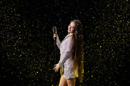Portrait of a beautiful singer with a vintage microphone against the background of falling snow. Young woman on an empty stage in the rays of yellow studio light close up.の写真素材