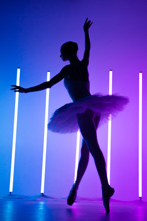 Young graceful ballerina in pointe shoes and a white tutu demonstrates her dancing skills. A beautiful classical ballet dancer against the backdrop of bright neon lights in a dark studio. Silhouette.の写真素材