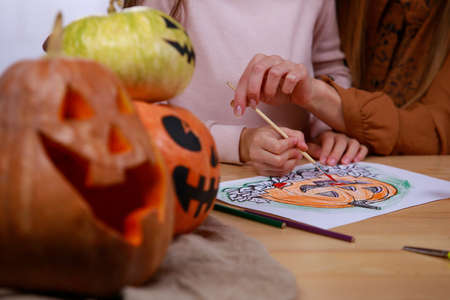 The family is preparing for Halloween and drawing pumpkins on paper with gouache, sitting at the table. Close up of hands of mom and daughter.の写真素材