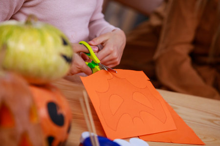 Preparing for Halloween. Close up of a girls hands cutting out a bat shaped mask on orange paper. Girl at the table with Halloween pumpkins, the foreground is blurred.の写真素材