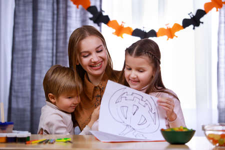 The family preparing for Halloween. Mom, daughter and son are sitting at the table in a room decorated for the holiday, laughing, hugging and examining the pictures with pumpkins. Close up.の写真素材