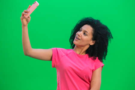 Portrait of young female African American making selfie on mobile phone. Black woman with curly hair in pink tshirt poses on green screen in the studio. Close up.の写真素材