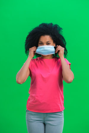 Portrait of young female African American looking at the camera and puts on medical mask. Black woman with curly hair in pink tshirt poses on green screen in the studio. Close up.の写真素材