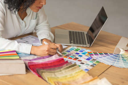 Portrait of a young African American woman looking through the color palette and samples of fabrics for a design project. Female designer in white blouse sits at a table in light office. Close up.の写真素材