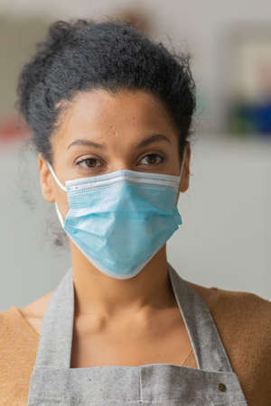 Pretty African American woman in medical mask. Young female mixed race posing in an apron against the backdrop of a light room. Close up.の写真素材