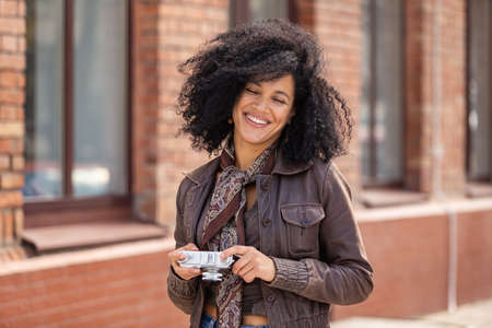 Stylish young African American woman smiling happily and holding retro camera. Brunette with curly hair in brown leather jacket poses against background of blurred brick building. Close up.の写真素材