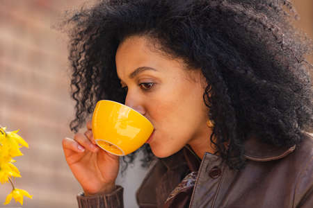 Portrait of stylish young African American woman enjoys aroma and taste of hot coffee in yellow cup. Brunette in brown leather jacket sitting at a table in a cozy cafe on the street. Close up.の写真素材