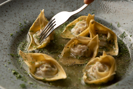 Fork pierces fresh wonton dumplings with meat presented on round gray plate close up. Restaurant serving food, Chinese traditional dumplings sprinkled with dill. Chinese cuisine. Asian food dim sum.の写真素材