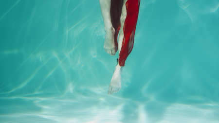 Underwater shot of woman dancing in red chiffon costume. Female dancer gracefully moves legs in blue water column with glare. Woman subaquatic shot, close up in slow motion.の写真素材