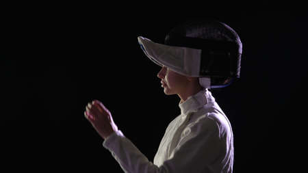 Side view of young female fencer putting protective helmet and holding fencing rapier in her hands....