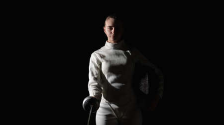 Portrait of a young woman fencer saluting with rapier in greeting. An athlete in a white uniform poses in a dark studio on a black background with a staged light. Slow motion. Close up.の写真素材