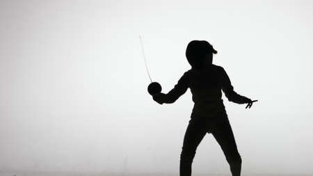 Silhouette of young woman swordsman throwing attacking blows with sword. Sports training in fencing school. Girl athlete posing in dark studio on a white background in spot of spotlight. Slow motion.の写真素材
