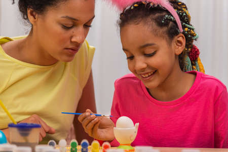 Cute little girl with funny bunny ears decorates eggs with brush and paints. African American mom...