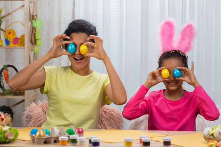 Mom and daughter with funny bunny ears laugh and have fun with colored eggs. African American woman and little girl sitting at table in decorated room at home. Happy easter. Close up.の写真素材