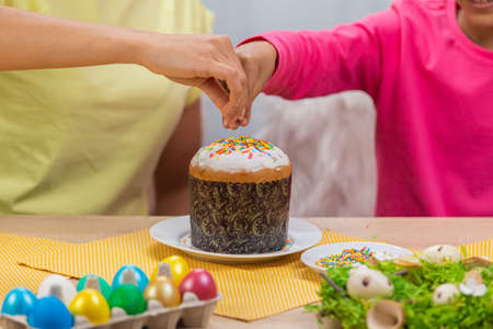 Mom and daughter decorate Easter cake with sweet candies. African American woman and little girl are sitting at table in festively decorated room. Happy easter. Close up.の写真素材