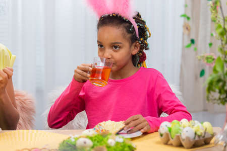 Mom and daughter with funny bunny ears are drinking tea with Easter cake. African American woman and...