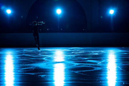 Young female figure skater is performing womans single skating choreography on ice rink. Woman practicing skills on ice arena against background of soft blue light and spotlights. Silhouette.の写真素材