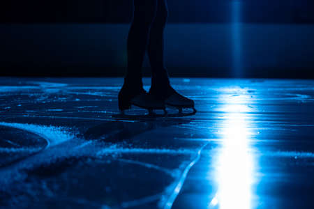Dark silhouette of female legs in figure skating skates sliding on ice arena. Young woman trains on dark ice rink with blue light. Shiny smooth surface of ice with scratches from skates. Close up.の写真素材