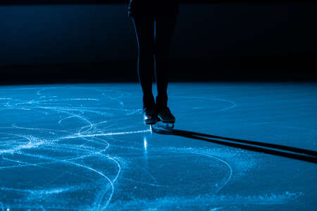 Dark silhouette of female legs in figure skating skates sliding on ice arena. Young woman trains on dark ice rink with blue light. Shiny smooth surface of ice with scratches from skates. Close up.の写真素材