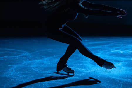 Figure skating lady is wearing black sportswear is skating on ice rink, training at night in the rays of blue light. Young woman preparing to competition, training rotation and slides skills. Close upの写真素材