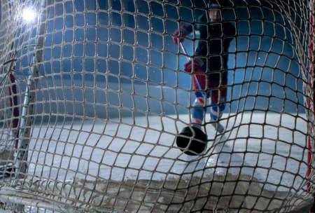 View from behind the net of the gate at a male hockey player scoring a goal. An athlete in uniform and skates plays hockey on a dark ice arena. Hockey player hits the puck with a stick. Close up.の写真素材