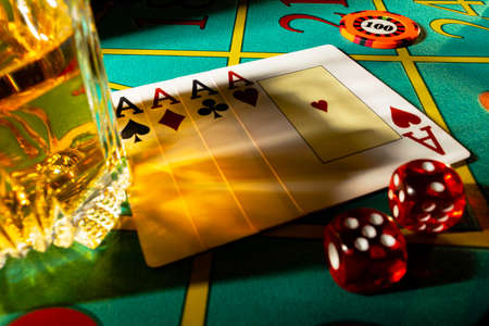 Gaming table in casino with glass of whiskey and set of four aces. Close up of a dark gambling poker table with booze, cards, dice and casino chips. The concept of gambling or poker and entertainment.の写真素材