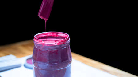 Pink paint flowing from brush into bank. Can of paint on table on black background. Process of preparing materials for dyeing paper tubes for weaving baskets. Creative work in workshop. Close up.の写真素材