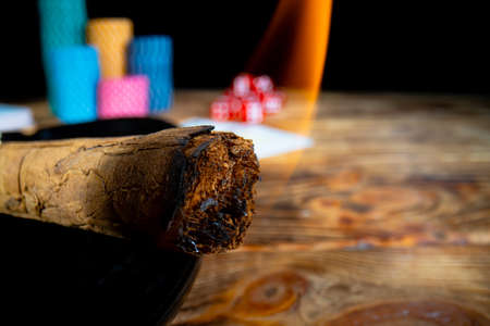 Smoking Cuban cigar against the backdrop of casino chips, cards and dice lying on wooden table. Burning Cuban cigar on an isolated black background in casino. Extra close up.の写真素材