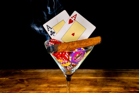 Burning cigar, cards, set of dice and casino chips in martini glass on black background. Two aces, chip denominated in one thousand and red dice on a wooden table. Gambling background. Close up.の写真素材