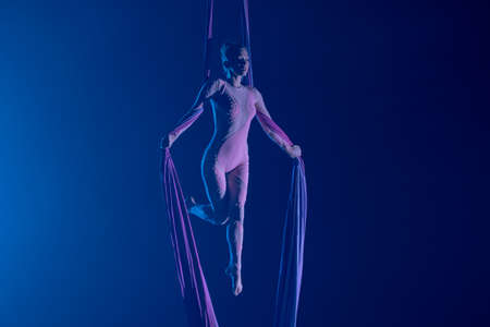 A young woman performing in a circus on aerial silk in the dark with blue light. A female equilibrist balancing on a height. Performance of aerial gymnast.の写真素材