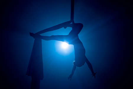 A young woman performing in a circus on aerial silk in the dark with blue light. Silhouette of a female equilibrist hanging upside down and demonstrating the twine. Performance of aerial gymnast.の写真素材