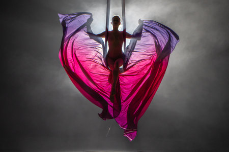 Silhouette of a young woman balancing on an airy silk ribbon and waving a cloth like butterfly wings. A female athlete performing in a circus at a height in the dark with backlight.の写真素材