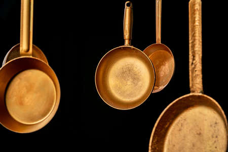 Set rustic copper fryingpans hanging on isolated black studio background. Round empty gold pans with handles. Kitchenware, utensils for cooking and frying. Restaurant interior background.の写真素材