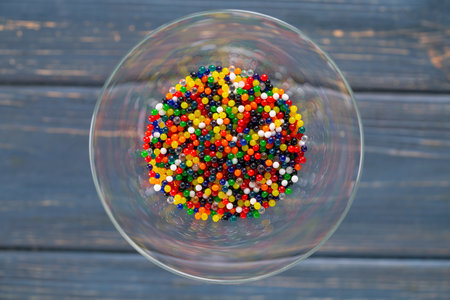 Top view of a martini glass with colored small balls. Bright red, blue, green, yellow and white particles in round glass on a gray wooden table. Festive screensaver. Wallpaper for birthday or holiday.の写真素材