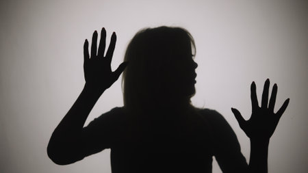 Black silhouette of a woman touching the glass with her palms and looking at the camera. The outline of a woman against the background of a window with white matte light. Close up.の写真素材