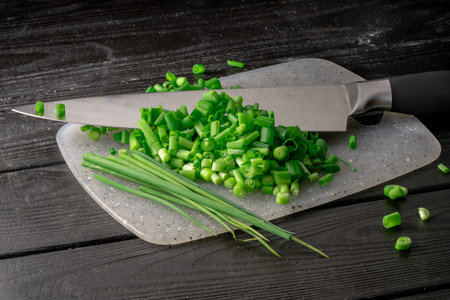 Chopped pieces and of green onion stalks next to a sharp metal knife on gray plastic board. Slices...