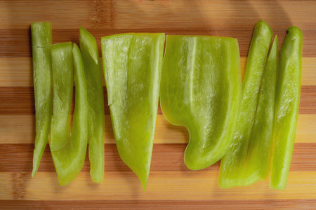 Pieces of raw green sweet pepper on striped brown wooden board. Slices of bell pepper with juicy pulp on the kitchen table. Chopped vegetables for making a fresh healthy salad. Close up. Top view.の写真素材