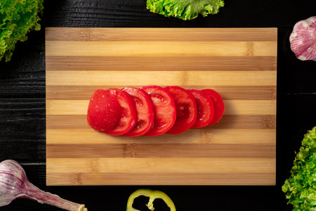 Top view of red ripe tomato with water drops is cutting into rings on a wooden kitchen board. Close up of sliced tomato, green lettuce, garlic and sweet pepper on black surface of wooden wet table.の写真素材