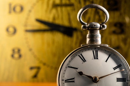 Silver antique pocket watch with Roman numerals and vintage table clock in blurry background. An old round white dial of pocket watch and yellow face of table clock with black hands. Close up.の写真素材