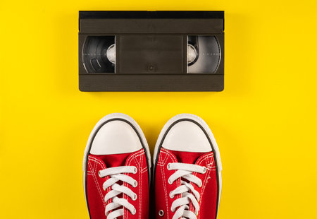 Black vintage videotape cassette and red white sneakers on yellow background. Plastic retro video cassette with analog magnetic tape from 90s and moccasins. Retro, vintage, nostalgia. Top viewの写真素材